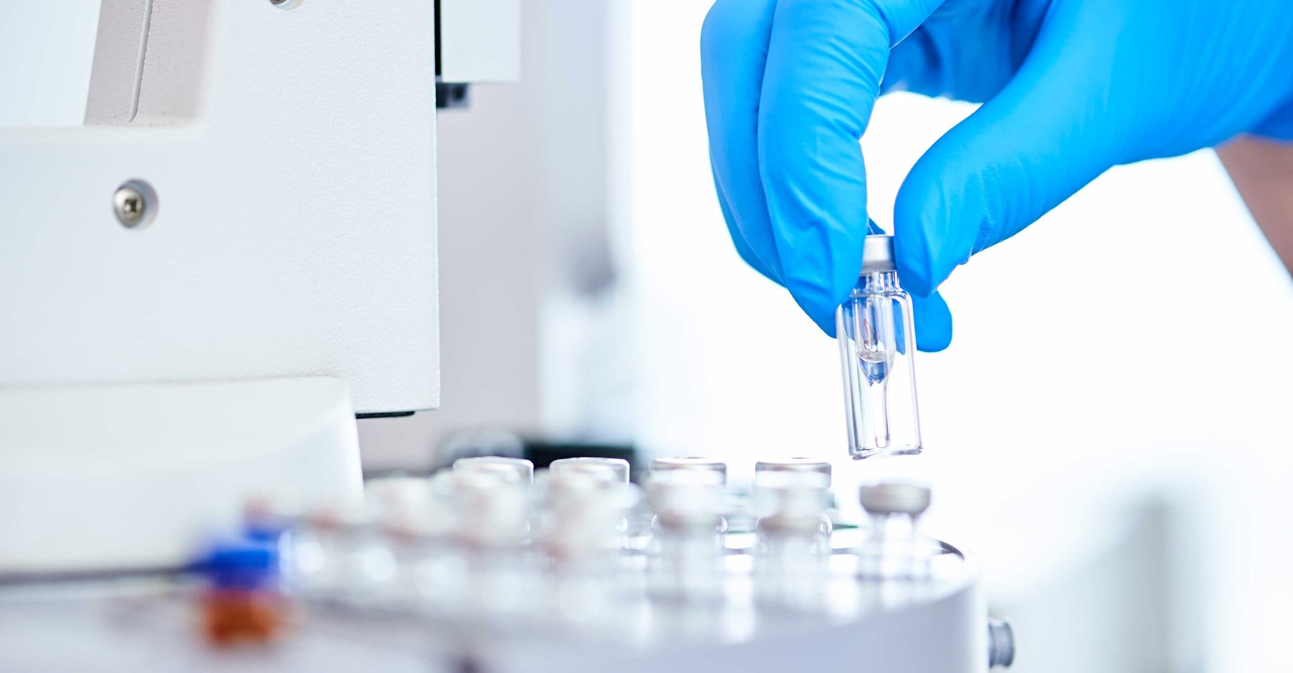 Eine Hand in einem blauen Handschuh hält ein kleines Glasfläschchen über eine Maschine im IHP-Labor.