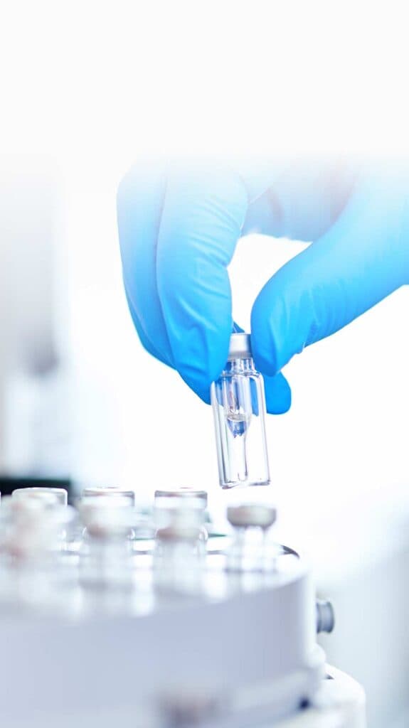 Eine Hand in einem blauen Handschuh hält ein kleines Glasfläschchen über eine Maschine im IHP-Labor.